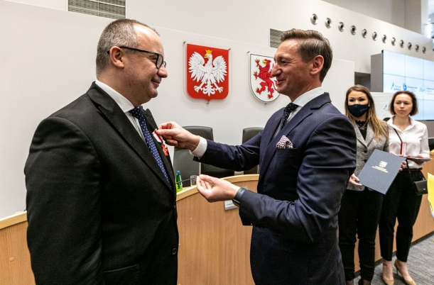 Dwa mężczyźni w garniturach stoją w dużej sali, nieco z tyłu widać dwie kobiety, stół prezydialny, a na ścienie godło państwowe i herb województwa zachodniopomorskiego. Jeden z mężczyzn przypina drugiemu, w klapę marynarki, odznaczenie