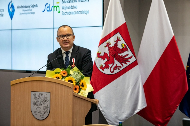 Mężczyna w okularach i ciemnym garniturze stoi przy mównicy. Na blacie, pod mikrofonem, leży bukiet kwiatów. Obok - stojak z biało-czerwonymi flagami. Na ekranie w tle wyświetlają się napisy, m.in. Młodzieżowy Sejmik Województwa Zachodniopomorskiego.