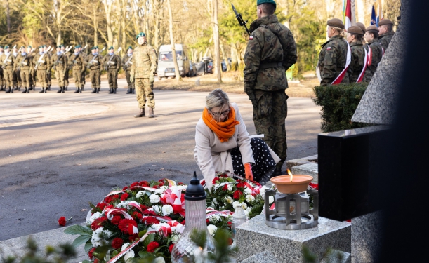 Kobieta, przyklękając, składa wiązankę kwiatów z biało-czerwoną wstęgą. Przy pomniku widać również inne wiązanki i wieńce oraz żołnierza pełniącego asystę honorową.