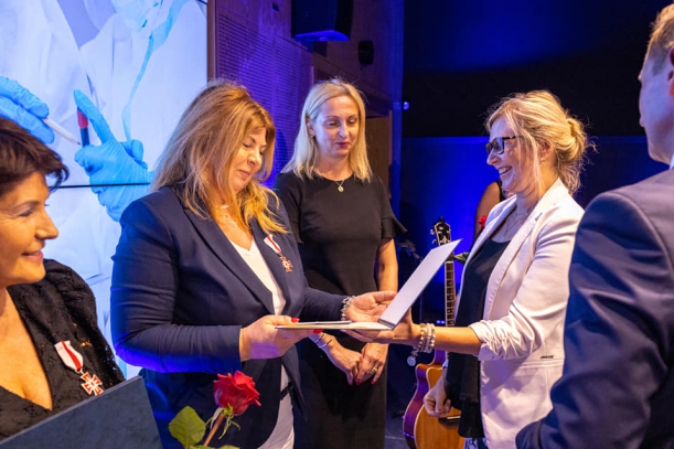 Członkini Zarządu Województwa wręcza odznakę kobiecie - dyr. szpitala w Kołobrzegu, w tle osoby i baner. 