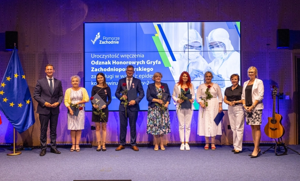 Grupa osób wyróżniona odznakami, nagrodzeni stoją na scenie na tle baneru nawiązująćego do COViD-19