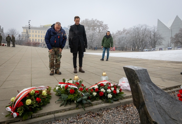 Marszałek Olgierd Geblewicz i prezes fundacji Wielka Orkiestra Świątecznej Pomocy Jerzy Owsiak przed pomnikiem Anioła Wolności w 52. rocznicę Szczecińskiego Grudnia. Stojąc na baczność, schylają głowy. Przed nimi - znicze i wiązanki kwiatów.