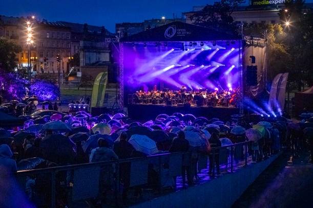Koncert "Sierpniowe Przełomy" na placu Solidarności w Szczecinie, 27 sierpnia 2023 r. Duża plenerowa scena oświetlona na fioletowo. Na scenie - kilkudziesięciu muzyków i dyrygent. Na pierwszym planie - publiczność, ludzie stoją pod parasolami.