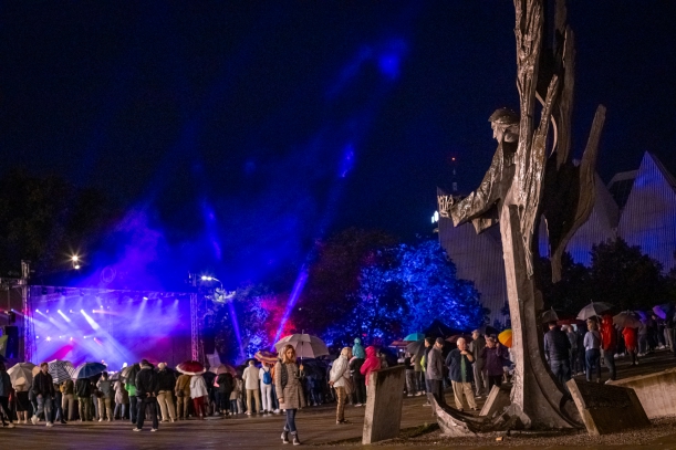 Koncert "Sierpniowe Przełomy" na placu Solidarności w Szczecinie, 27 sierpnia 2023 r. Na pierwszym planie - pomnik Anioła Wolności; przy nim kobieta z parasolem, patrząca na tablicę z nazwiskami Ofiar Grudnia'70. W tle - oświetlona scena i kilkaset osób.