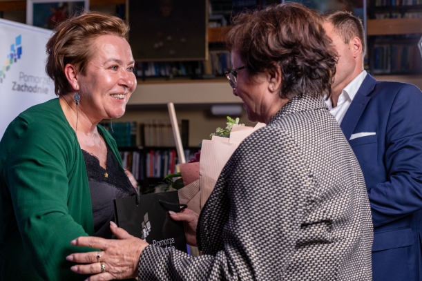 Spotkanie "Sydonia i Gryfici" (z cyklu "Otwarta społeczność") w Książnicy Pomorskiej. Przewodnicząca Sejmiku Województwa Zachodniopomorskiego Teresa Kalina wręcza torebkę z upominkiem pisarce Elżbiecie Cherezińskiej. Obok stoi mężczyzna z kwiatami.