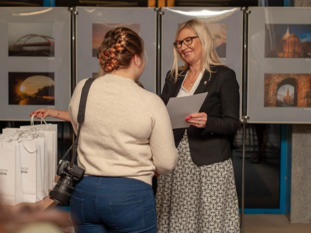 Wernisaż wystawy "Otwarte Pomorze Zachodnie. Oczami Imigrantów" w Książnicy Pomorskiej. Członkini Zarządu Województwa Anna Bańkowska składa gratulacje jednej z autorek zdjęć. Wręcza jej dyplom. Dziewczyna stoi z aparatem na pasku na lewym ramieniu.