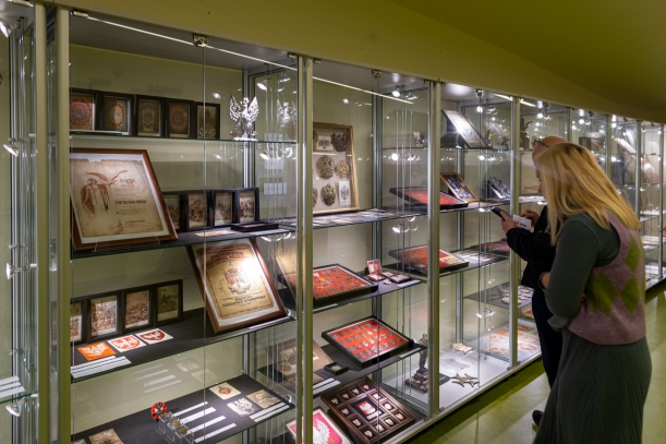 Oświetlone gabloty wystawy "Polskie orły". Na półkach ramki z wojskowymi przypinkami na czerwonym suknie, zdjęcia, grafiki z polskim godłem. Przy gablotach stoją kobieta i mężczyzna.