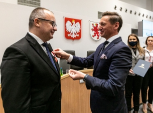 Dwa mężczyźni w garniturach stoją w dużej sali, nieco z tyłu widać dwie kobiety, stół prezydialny, a na ścienie godło państwowe i herb województwa zachodniopomorskiego. Jeden z mężczyzn przypina drugiemu, w klapę marynarki, odznaczenie